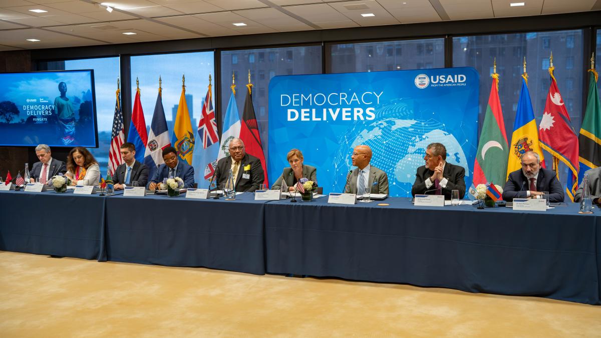 Fijian President Ratu Wiliame Katonivere, wearing a dark suit and gold tie, speaks with USAID Administrator Samantha Power, dressed in a light gray suit, at a formal roundtable, at the “Democracy Delivers” event co-hosted by USAID and the Ford Foundation during the UN General Assembly in New York. Various national flags, including Fiji’s, are displayed in the background.