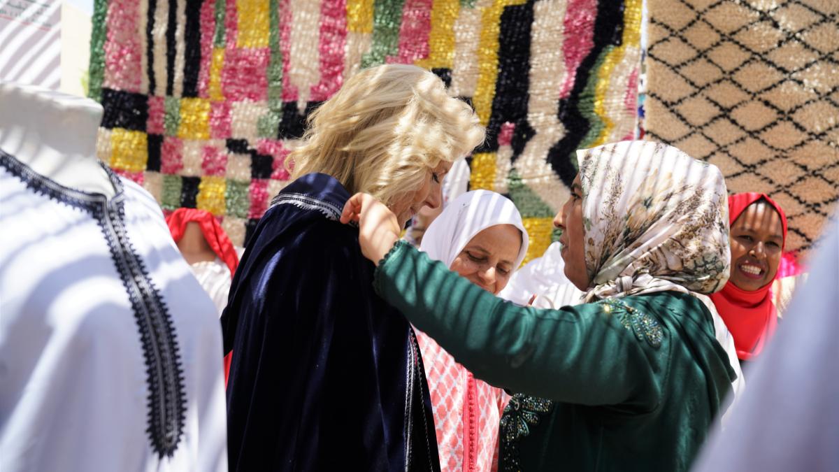 A woman in green traditional Moroccan dress and a beige hijab, right, fastens a traditional blue cape over the shoulders of U.S. First Lady Dr. Jill Biden, left, in front of Moroccan rugs that hang in the background.