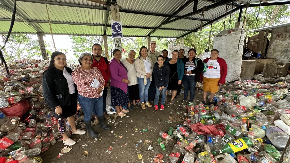 About 12 people pose for a group photo in a cleared, dirt space under an open tin-roof structure; they are surrounded by plastic bottles and other plastic products.