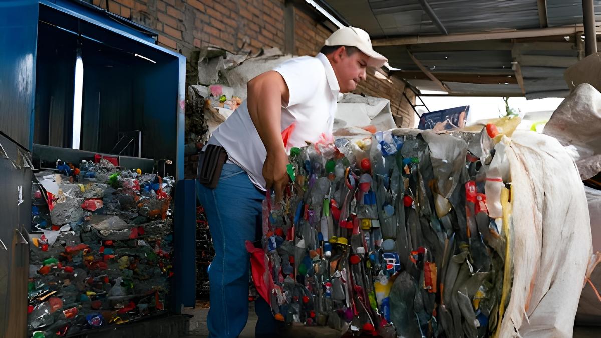  A man compiles compressed plastic bottles on a pallet; behind him, machinery compresses additional plastic waste.