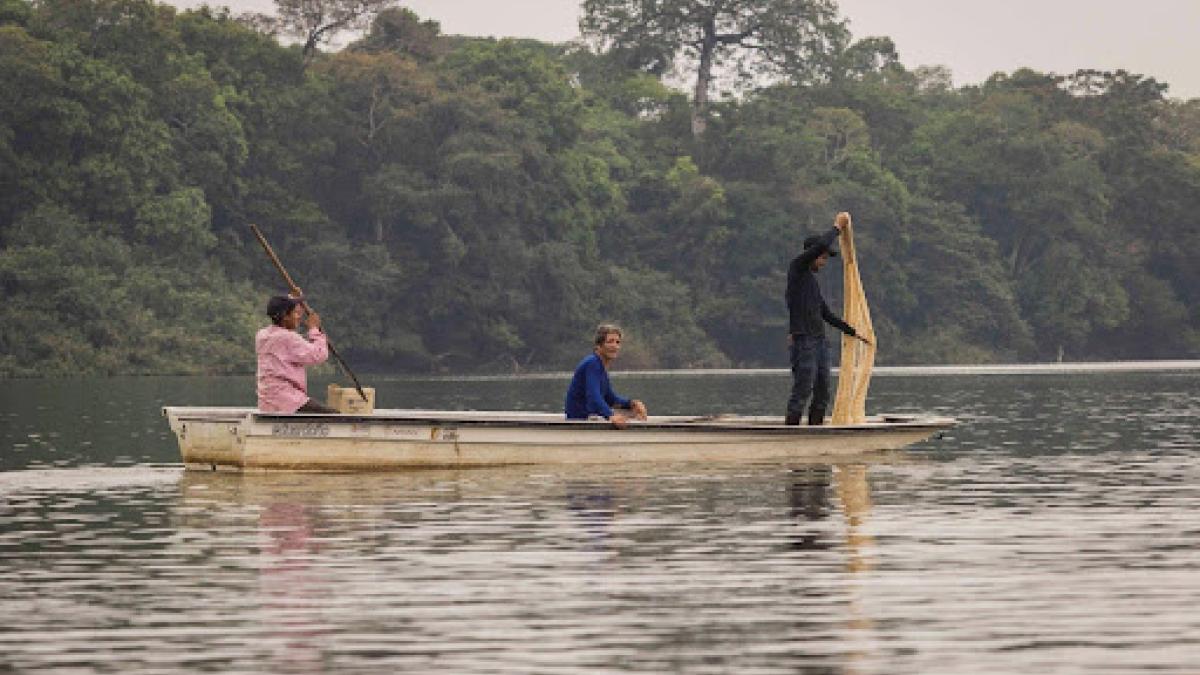 Three men ride in a small boat over a river, with one man in the front of the boat holding a fishing net, and the man in the back of the boat holding an oar. 