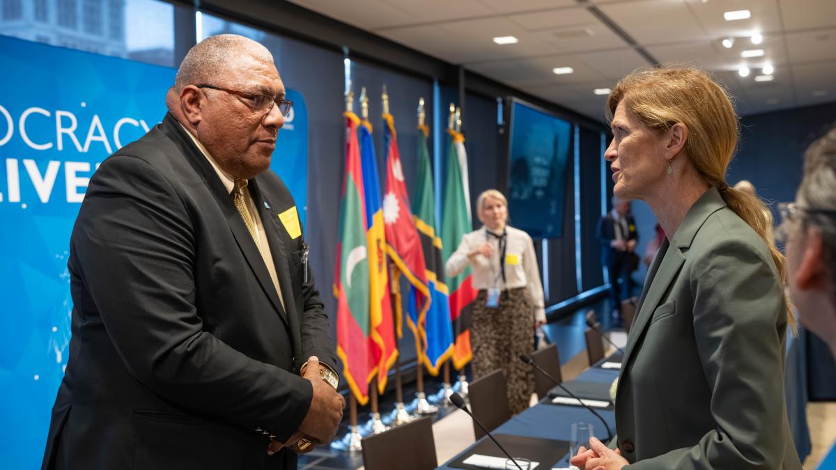 Fijian President Ratu Wiliame Katonivere, wearing a dark suit and gold tie, speaks with USAID Administrator Samantha Power, dressed in a light gray suit, at the “Democracy Delivers” event co-hosted by USAID and the Ford Foundation during the UN General Assembly in New York. Various national flags are displayed in the background.