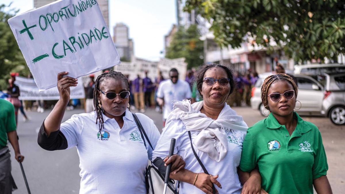 A woman wearing sunglasses holds a sign that reads, in Portuguese, “ + Oportunidades - Caridade” (or “more opportunities, less charity” in English) with her arms interlocked with two other women who are participating in a parade. 