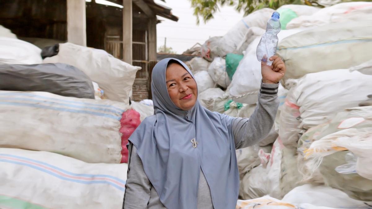 a woman stands in front of sorted debris, holding up a single-use plastic water bottle. 