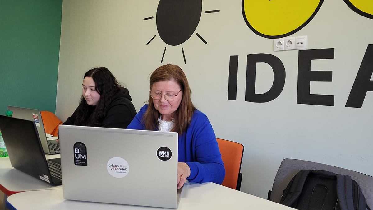 2 women work on their laptops at a table in front of a wall with lightbulb designs on it.