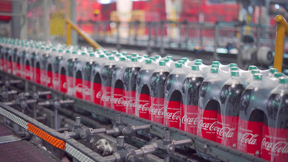 Large bottles of dark liquid labeled “Coca-Cola” with other markings in Spanish pass through machinery after bottling.