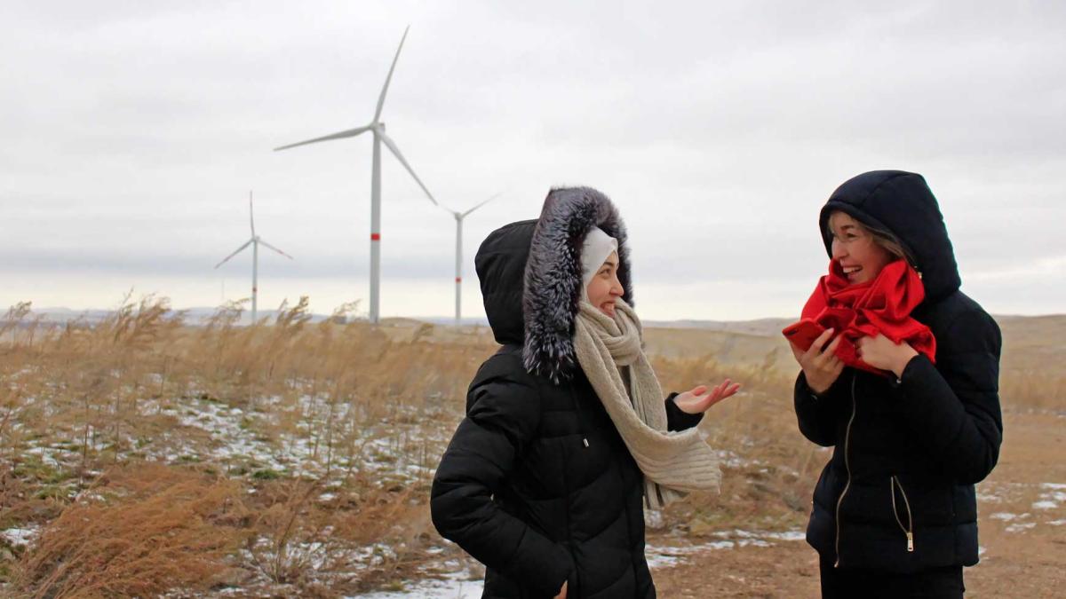 Two happy women in winter clothes chat in a dry amber field with wind turbines in the distance set against a gray sky