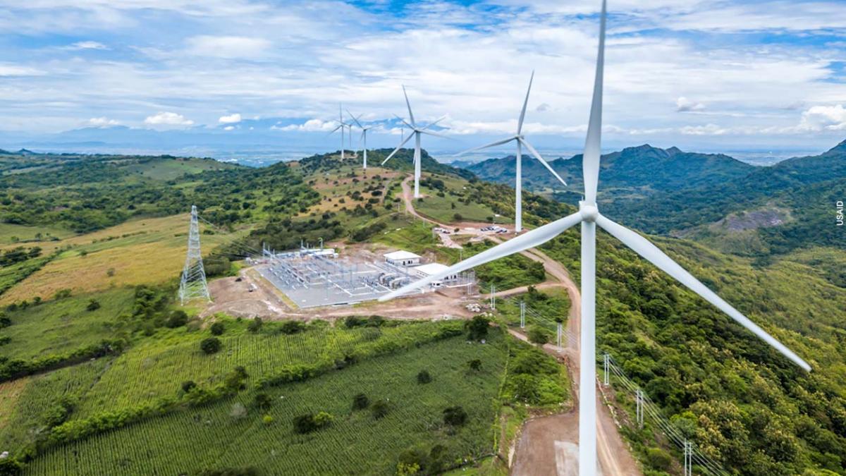 Aerial photo of a wind farm. A line of large wind turbines stretch into the distance along a green ridge against a blue sky filled with white clouds. The wind farm's power transmission facility can be seen in the center of the ridge.