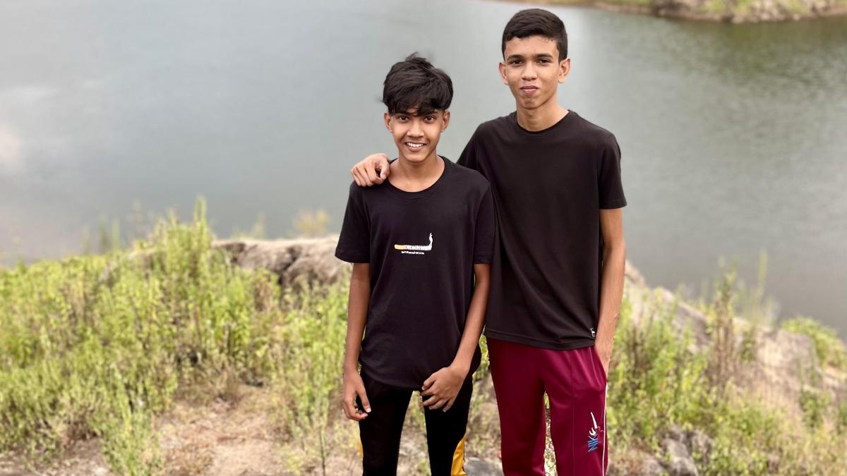 Two boys stand on a rock in front of a lake with a mountain in the background.