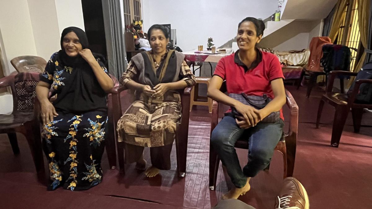 Three women seated on chairs in a bright room, sharing smiles and laughter as they chat together.