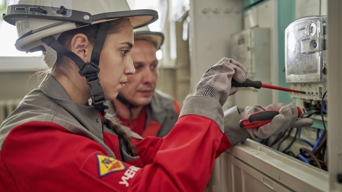 North Macedonia - A young woman in protect gear works intently on an electro fitter with her tools. Her training instructor, also in protective gear, stands next to her and observes. 