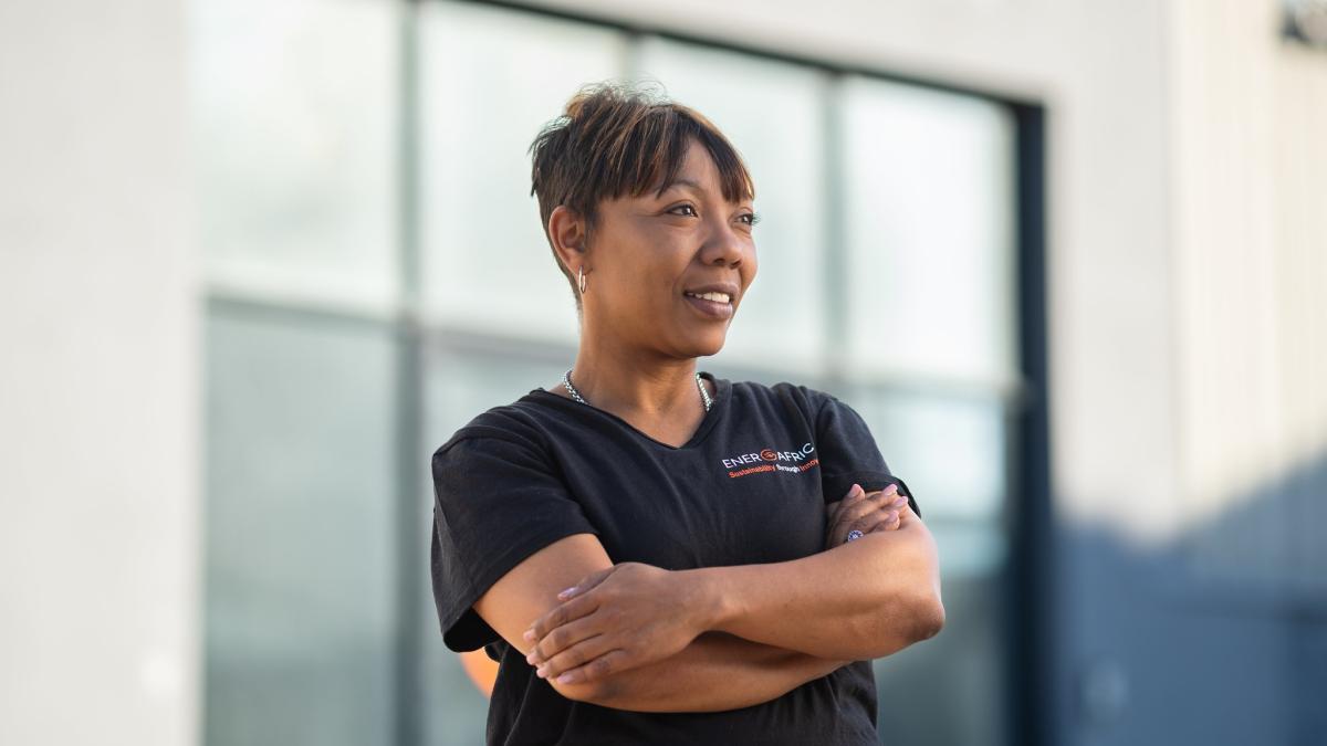 Woman wearing dark blue shirt with "ENERGAFRICA" logo looking to the side of the camera smiling. Windows of a building can be seen in the background. 