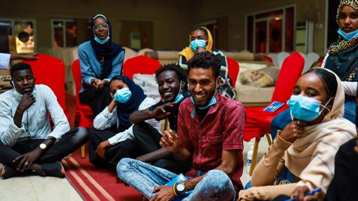 A group of young people with and without facemasks sitting together on a red carpet and chairs.