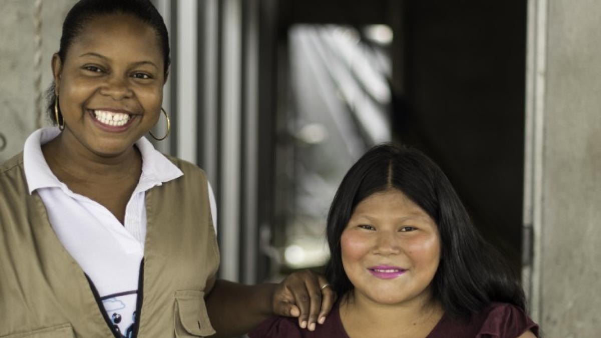 Two smiling women, one with her arm on the shoulder of the other, in front of an open doorway.