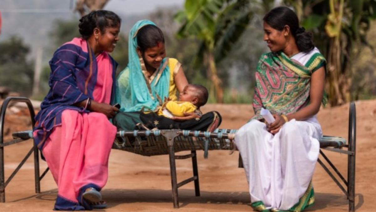 Three indigenous women sitting on a bench. One of the women is holding a baby and the other two are watching the child.