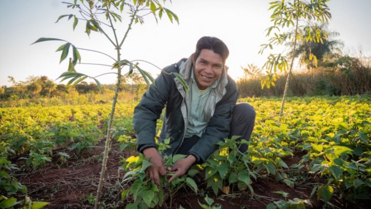 A smiling indigenous man wearing a gray jacket kneeling in a field.