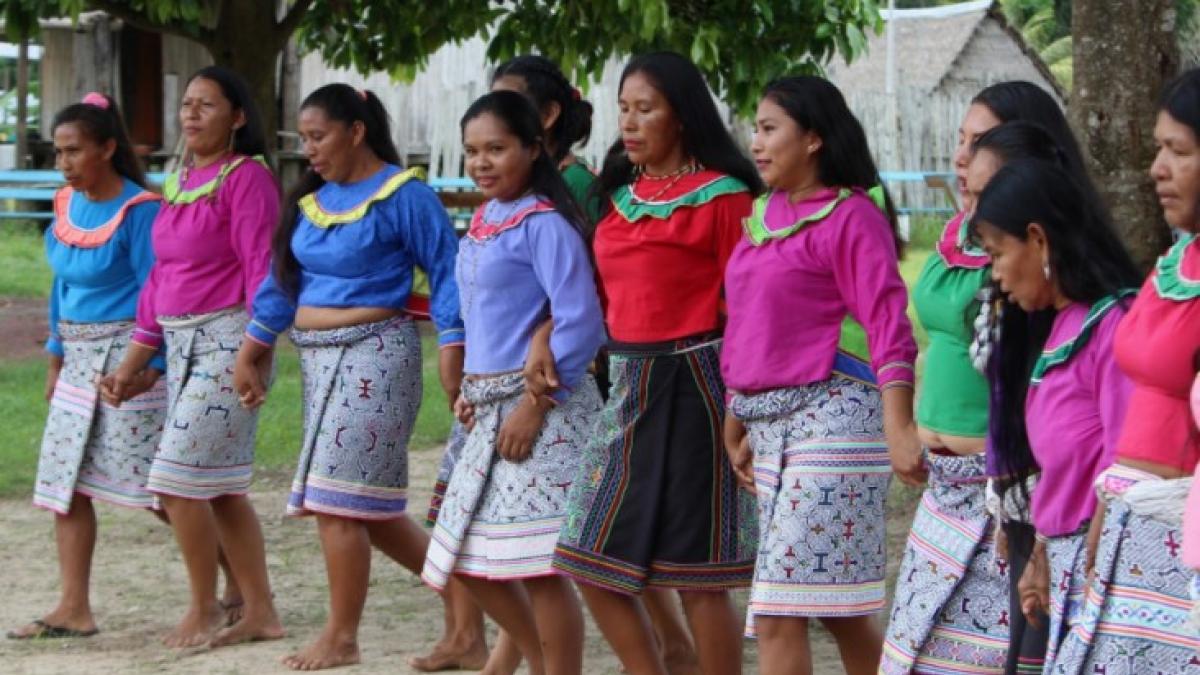 A group of women dancing outdoors. They are wearing colorful shirts and knee-length skirts with geometric patterns.