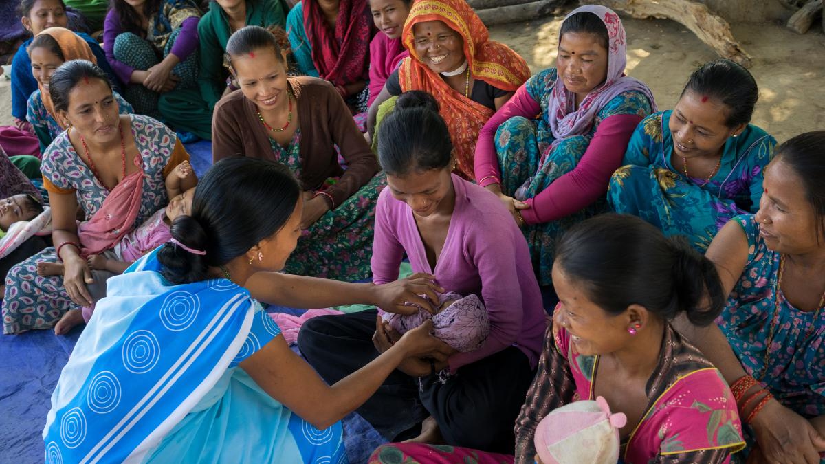 A group of women sit on the ground. Another woman crouching in front of them assists one with her baby.