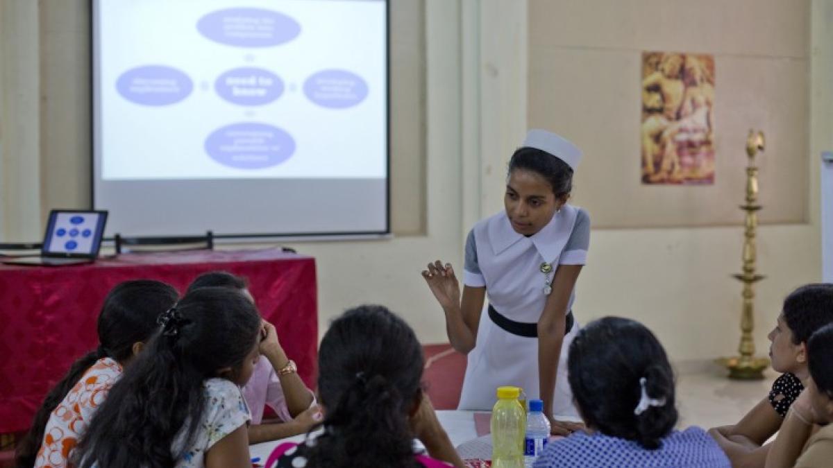 A woman wearing a nurse’s uniform leaning on a table and speaking to a group of people in a conference room.