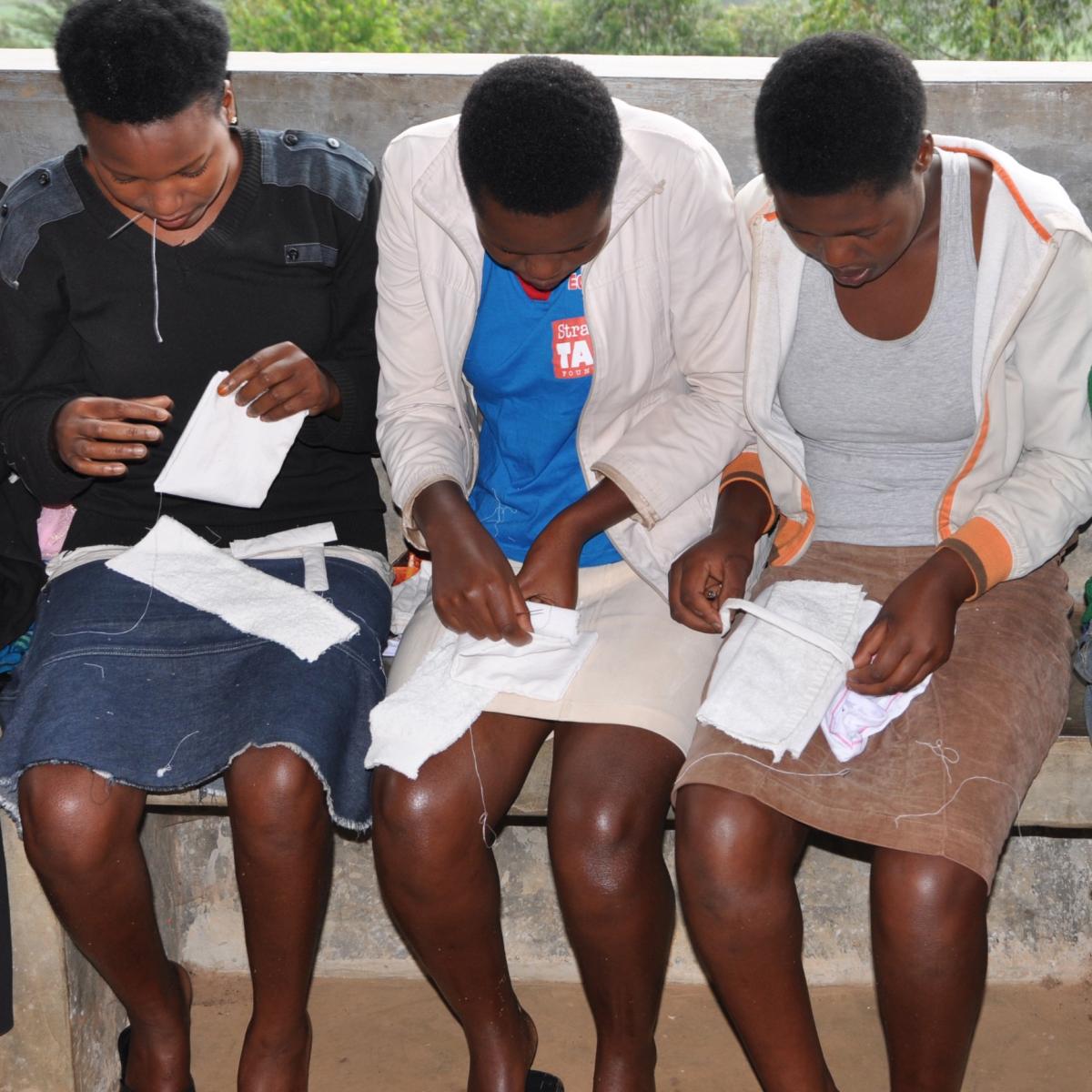 Three young women sitting in a row with materials to make menstrual pads in their laps