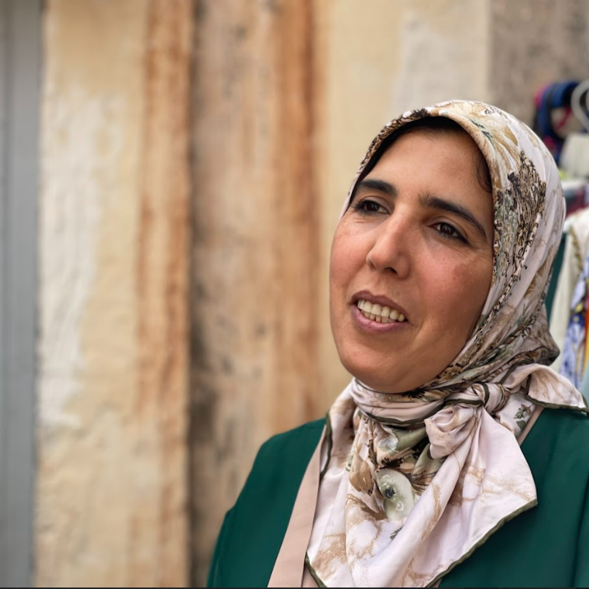 A Moroccan woman wearing a green dress and beige hijab smiles, behind her a rack of colorful clothing items