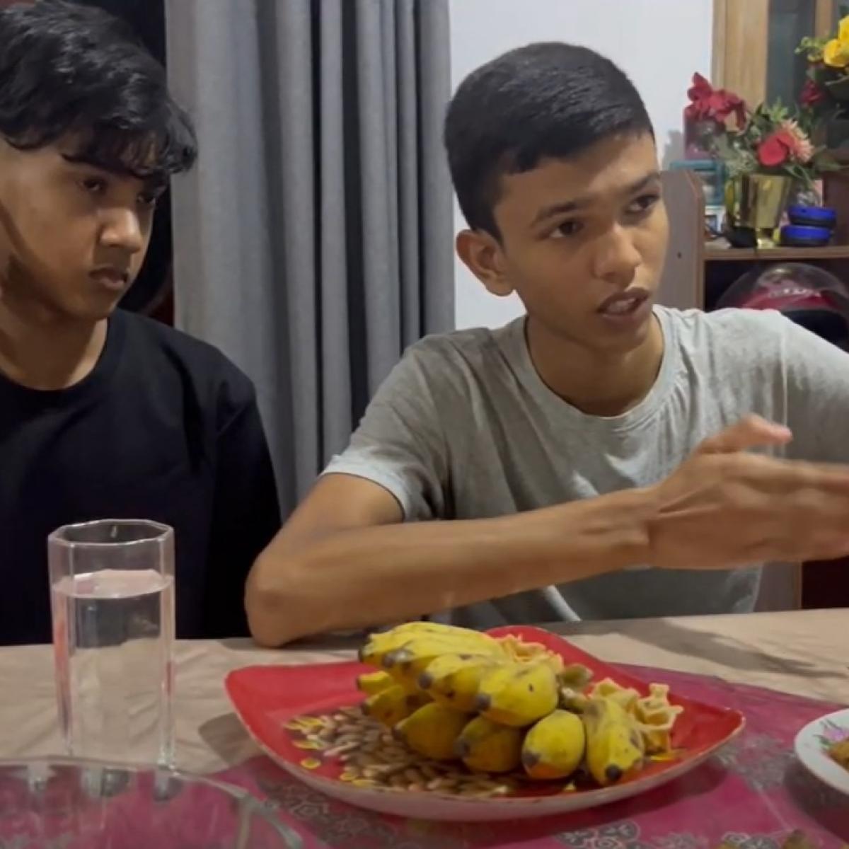 Two boys sit at a table with a spread of food, talking.