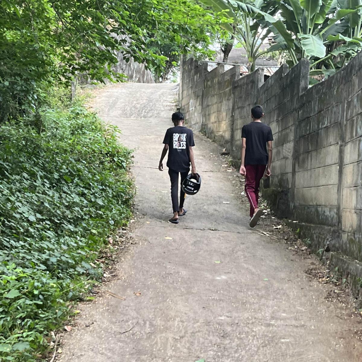 Two boys stroll uphill along a narrow path beside a tall wall.