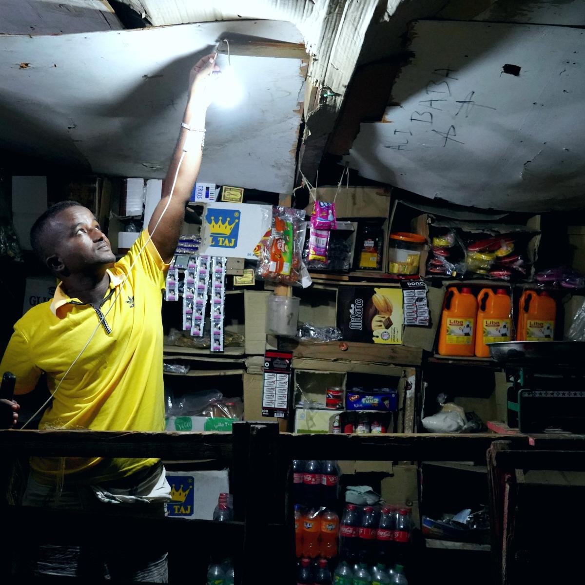 A shopkeeper lights his lamp to illuminate his shop utilizing mobile solar batteries. Kalaf Village, Djibouti.