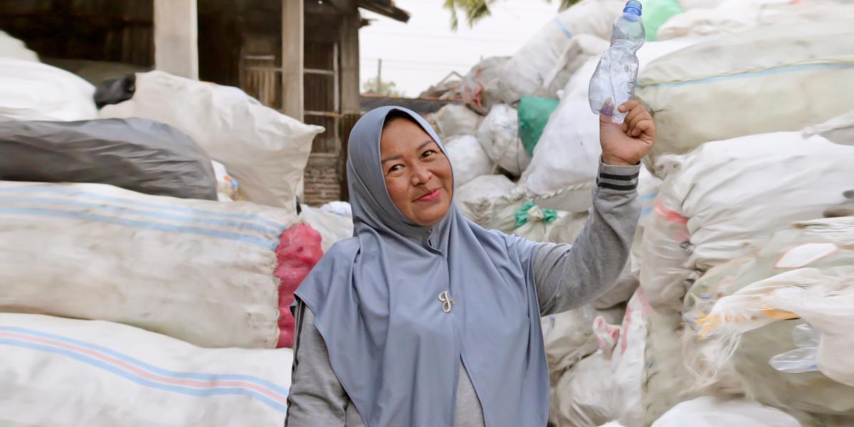 a woman stands in front of sorted debris, holding up a single-use plastic water bottle. 