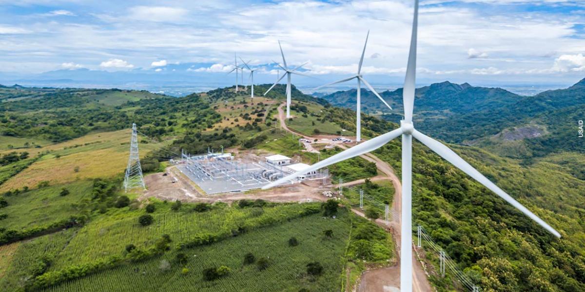 Aerial photo of a wind farm. A line of large wind turbines stretch into the distance along a green ridge against a blue sky filled with white clouds. The wind farm's power transmission facility can be seen in the center of the ridge.