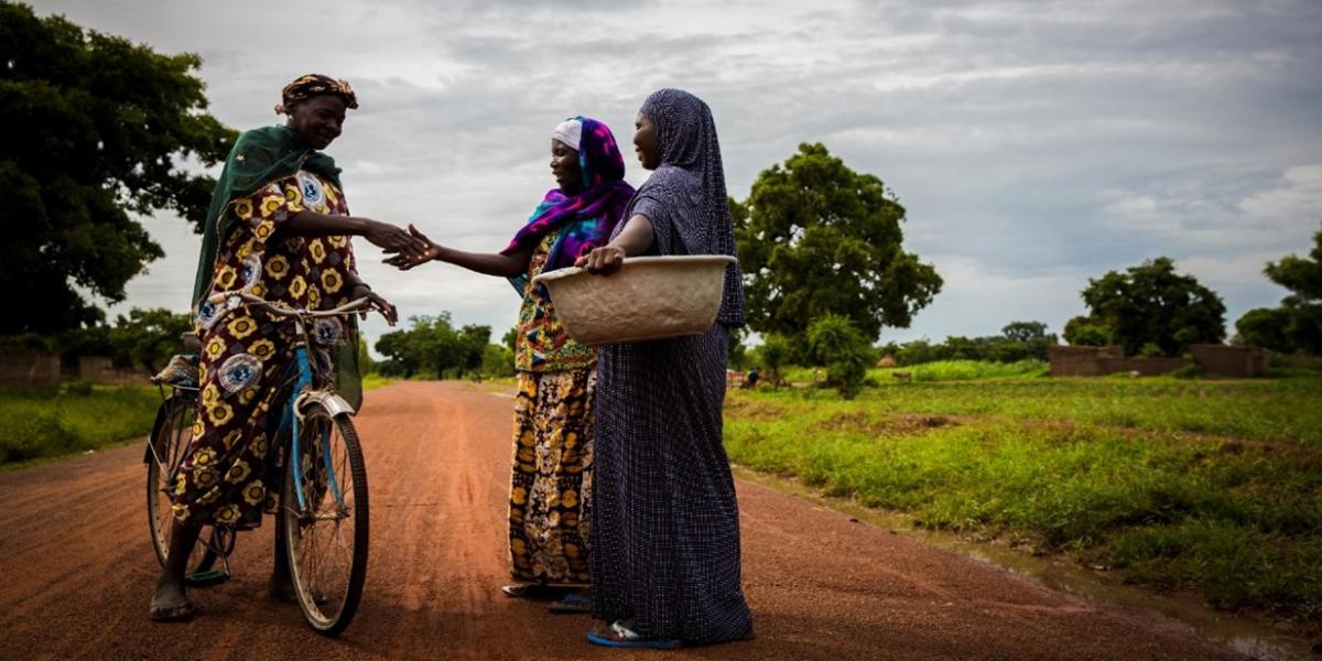 Woman shakes hands with another woman on bicycle, next to woman holding basket on a dirt road. 