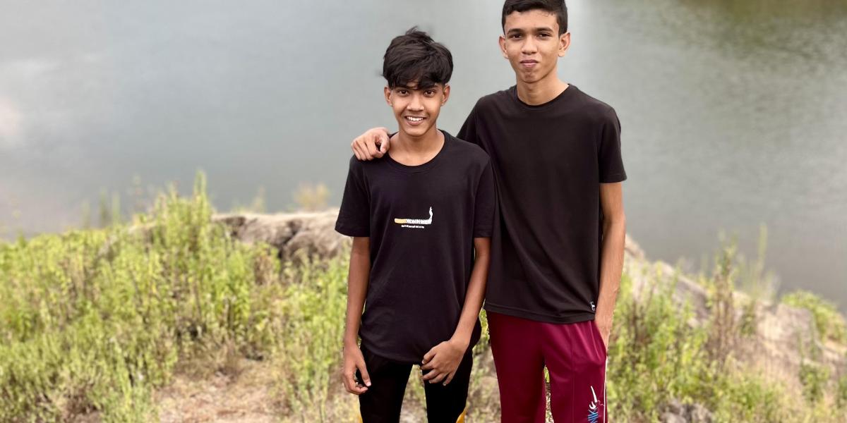 Two boys stand on a rock in front of a lake with a mountain in the background.