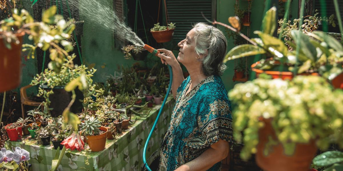 Centered in her lush nursery, Olga raises a hose to water her plants all around her. Potted succulents cover a tabletop and hang from wires.