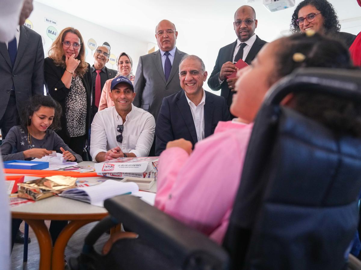 A group of adults in professional dress smile while looking at a child in a wheelchair as she laughs.