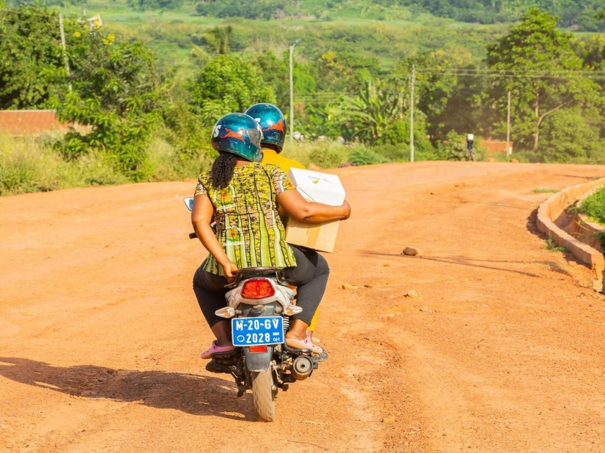 Two people on a motorcycle on a dirt road with the woman on the back carrying a box.