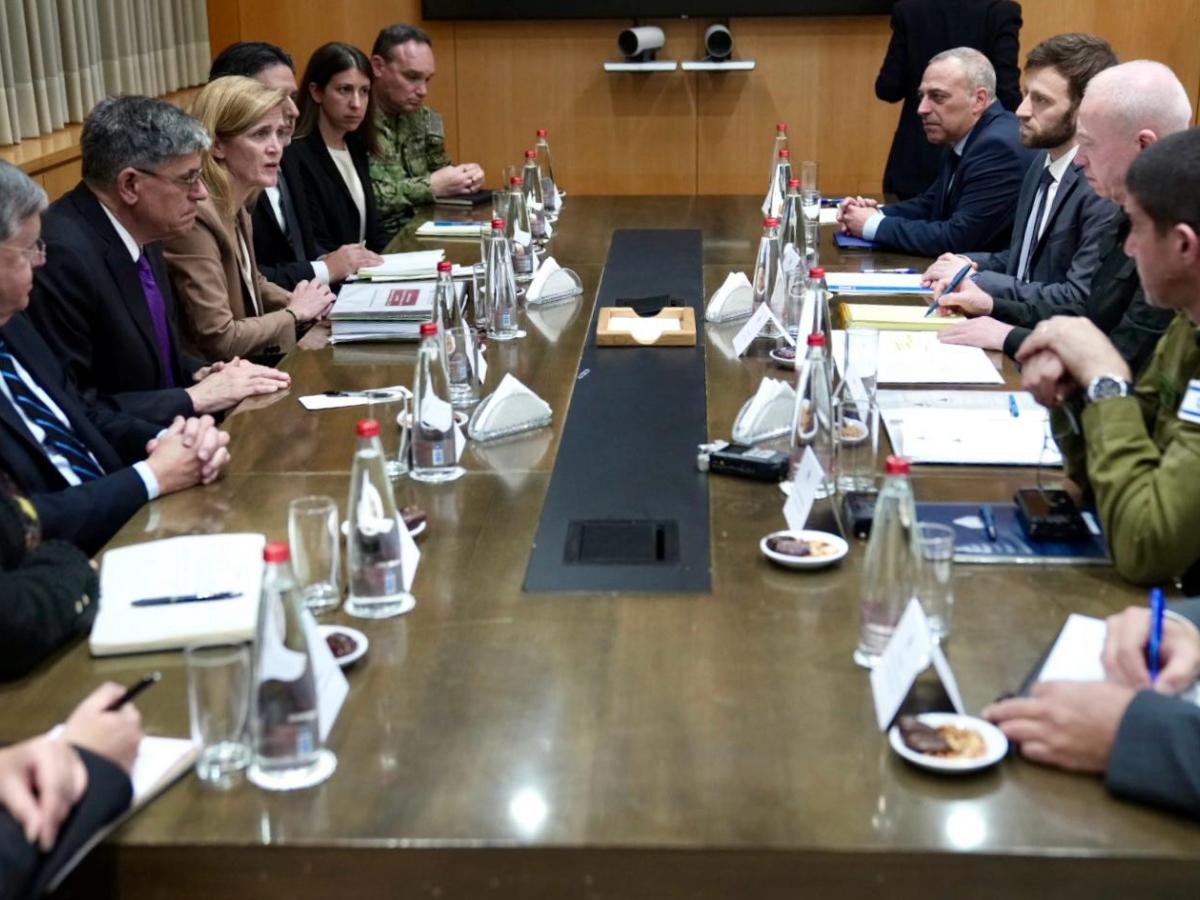 A group of men and women sitting around a long rectangle table in a meeting room engaged in discussion. 