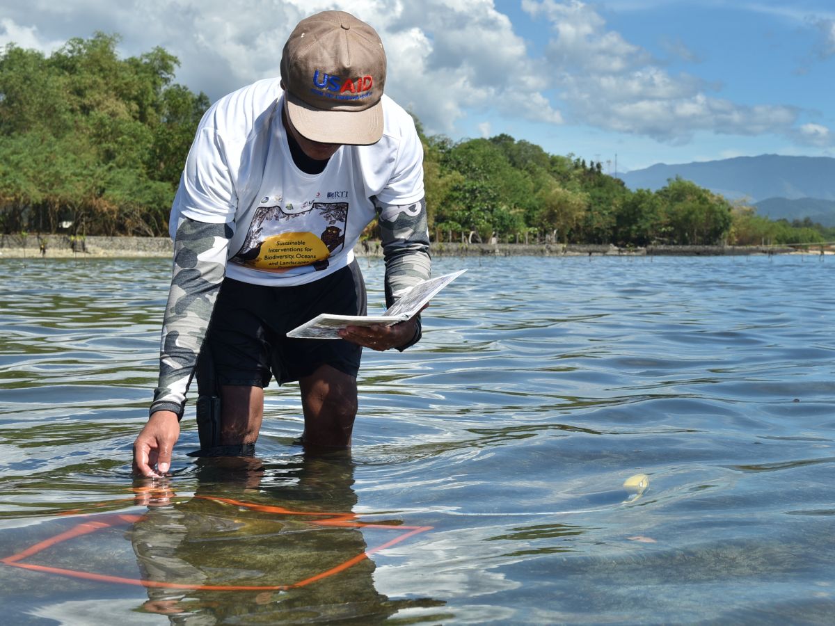 A man stands in shallow water with a book and tools used to complete testing.