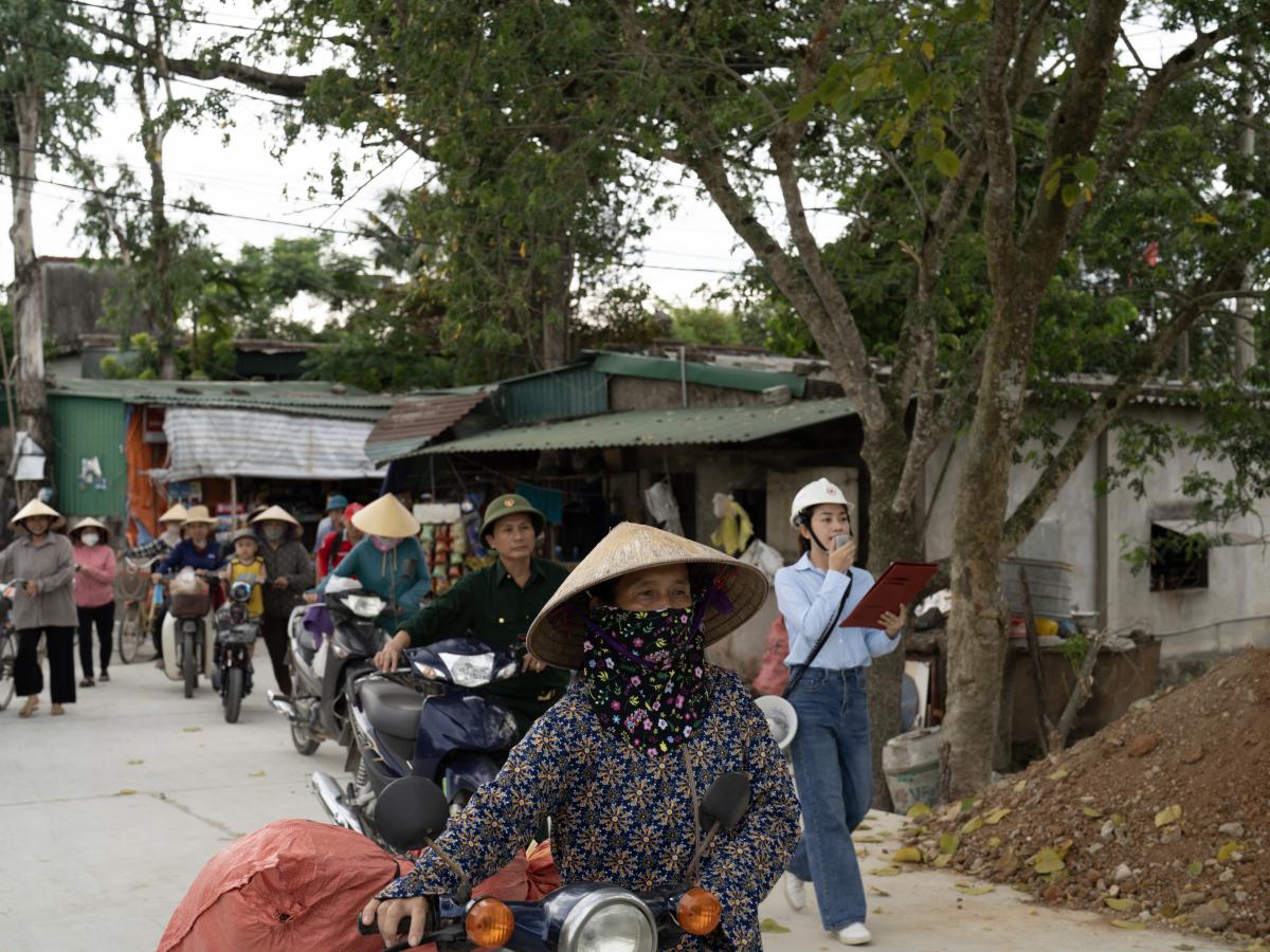 They stand patiently beside wheelbarrows, bicycles, and baskets loaded with supplies—rice, household items, and water—ready to participate in a USAID and Vietnam Red Cross Society (VNRC) disaster preparedness drill.