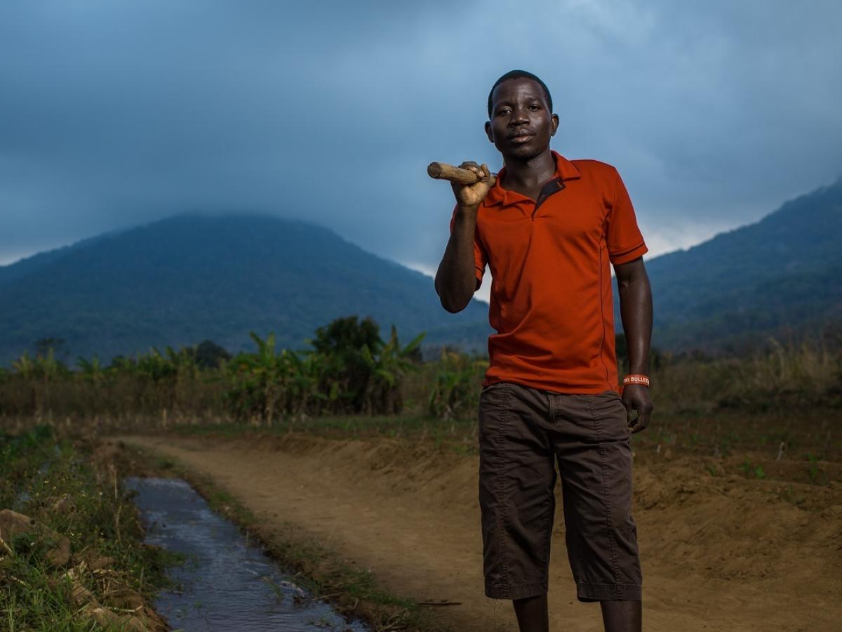 A man in an orange shirt and shorts stands with a tool over his shoulder next to an irrigation canal with mountains in the background