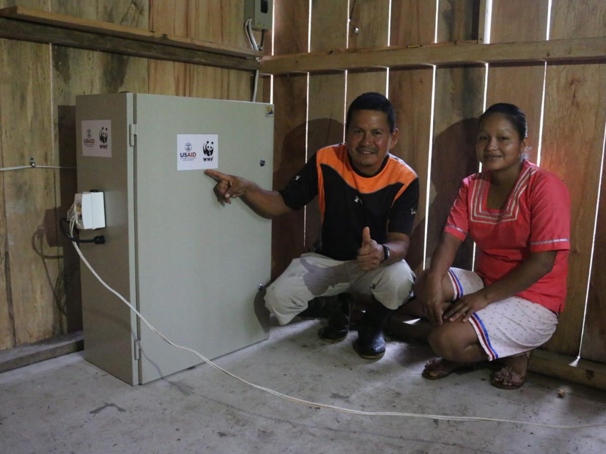 This picture shows two people standing in front of a piece of equipment with the WWF and USAID logo. The equipment contains the inverter and controls for the solar panel system. 