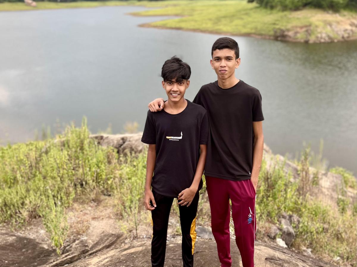 Two boys stand on a rock in front of a lake with a mountain in the background.