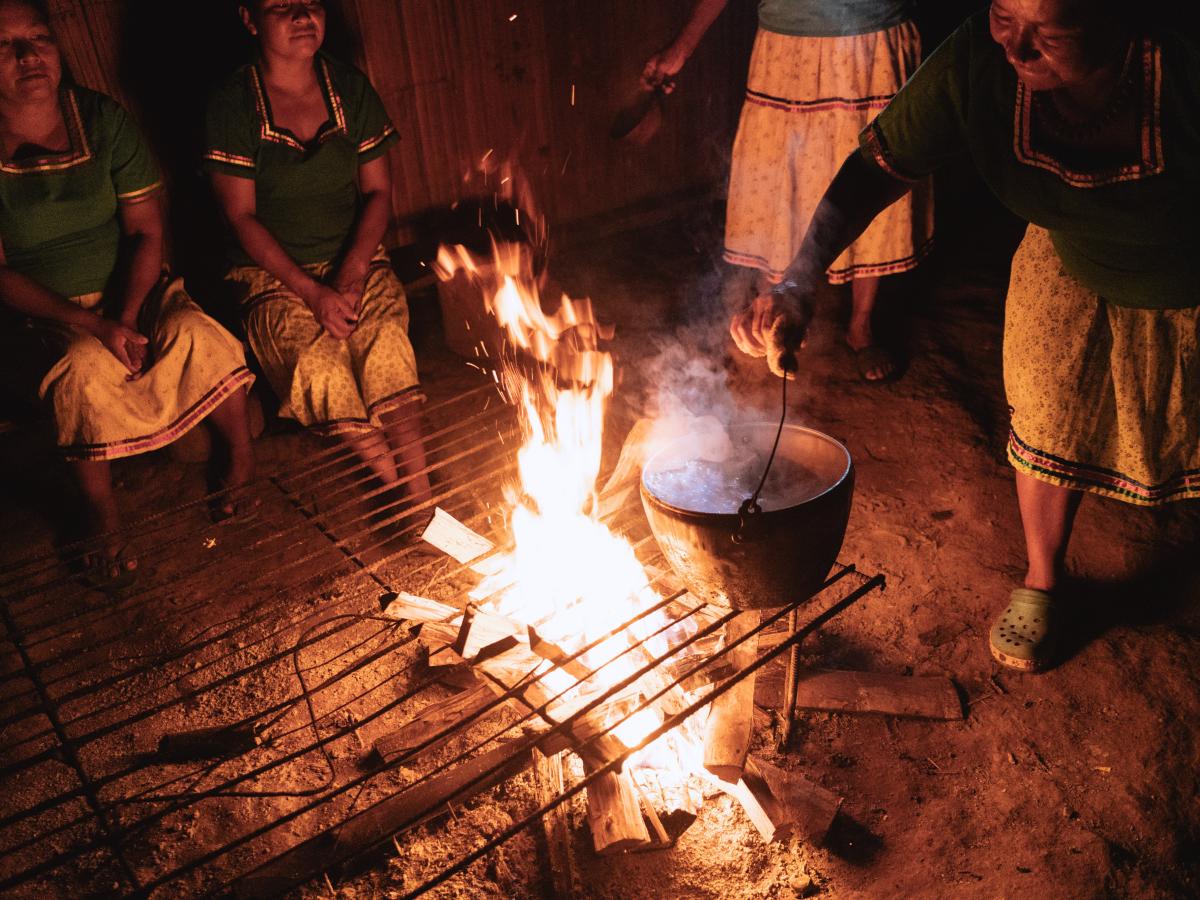 This picture shows four indigenous women sitting around a fire. One of them is placing a pot on the fire. They are wearing green shirts and white skirts. 