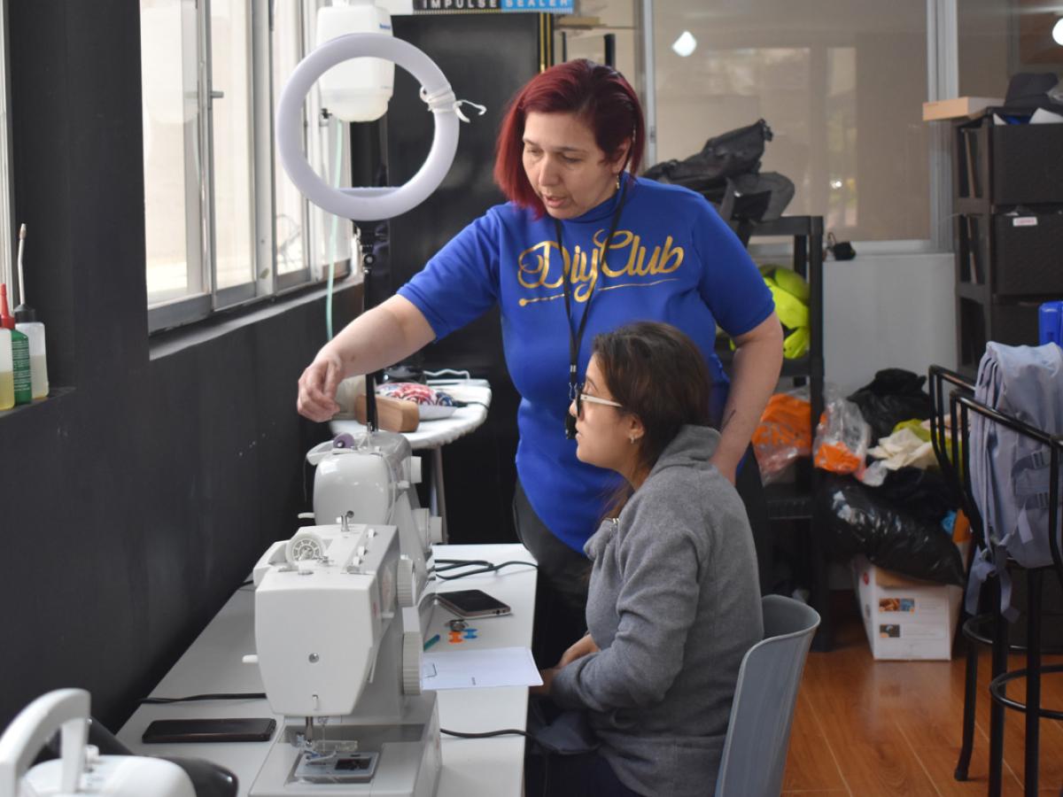 This picture shows Laura instructing a female student. They are using a sewing machine.