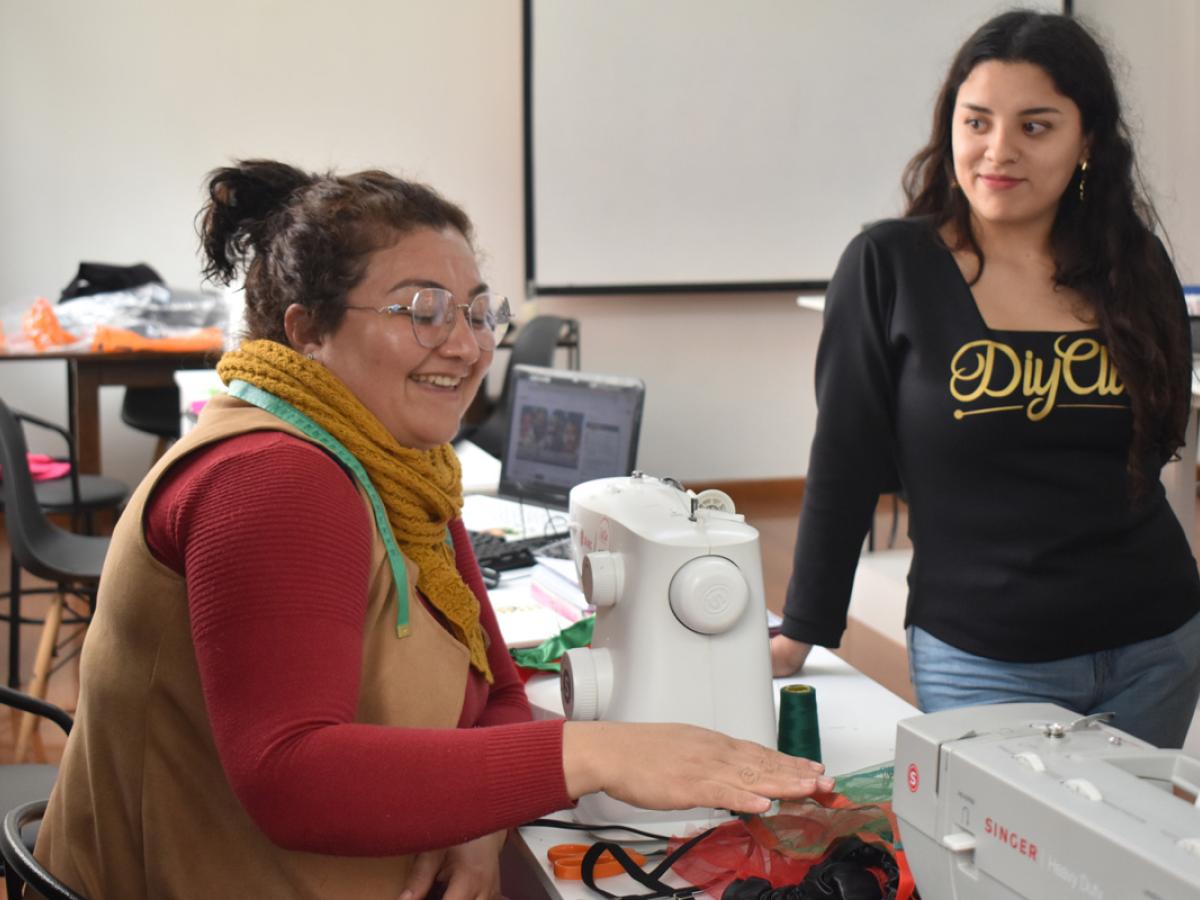 This picture shows Dani instructing a female student who is using a sewing machine. They are both in long-sleeved shirts. 