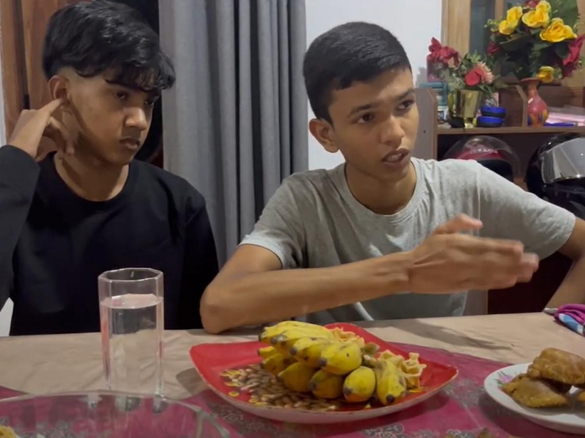 Two boys sit at a table with a spread of food, talking.