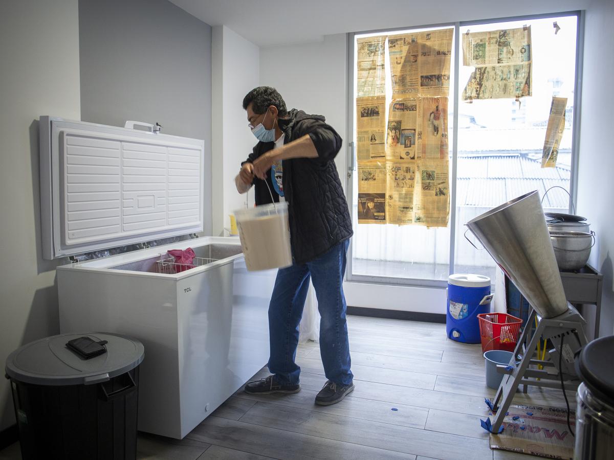 This picture shows Carlos Torres holding a bucket with his product, chicha. He seems to be storing it in a freezer. The room has a window. 