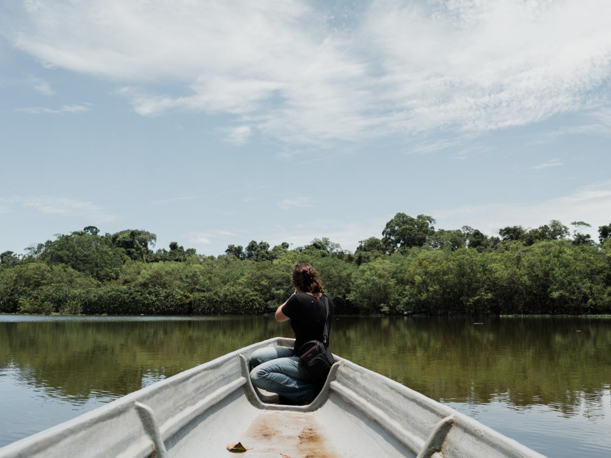 This picture shows the front part of a boat and a person sitting on it. The person is showing her back while he/she observes the river the boat is on. 