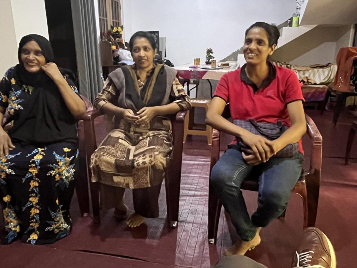 Three women seated on chairs in a bright room, sharing smiles and laughter as they chat together.