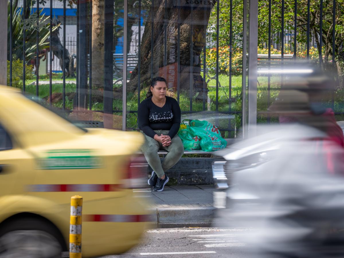 This picture shows Ariana, a female and main character in the story. She is sitting at a bus stop in the city while cars are going by in front of her. There are bags with groceries next to her. 