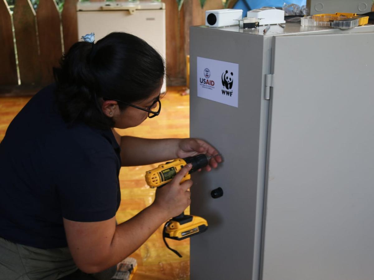 This picture shows a person, likely a woman, drilling something on a piece of equipment. The equipment has USAID and WWF stickers on it. There are more tools on top. 